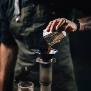 Barista pouring coffee into aeropress coffee maker. Barista with tattooed arms wearing dark uniform.